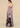 A woman modeling a strapless pleated maxi dress from the back, featuring a lavender bodice and a large abstract black, brown, and pink print on the skirt, set against a solid beige studio background.