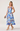 A woman modeling a sleeveless midi dress with a blue and purple floral print on a white background, holding a small clutch and wearing strappy high heels against a light gray studio backdrop.