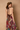 A woman shown from the back wearing a sleeveless brown halter dress with a tied back and a tiered multicolor skirt featuring a patchwork print in purple, red, green, and gold, posed against a textured beige wall.