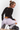 A woman crouches in a studio wearing a black long-sleeve textured crop top, light lavender leggings, gray socks, and black sneakers, with a black bag on the floor beside her.