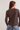 A woman shown from the back wearing a dark brown ribbed long-sleeve top with a small rectangular patch detail on the upper back, paired with blue jeans against a plain light background.