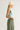 A woman in profile wearing a light blue ribbed tank top, an off-white knit beanie, and an olive green skirt, photographed against a plain light gray studio background.
