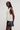 A model wearing a cream-colored, sleeveless open-knit vest with a side cutout and decorative buttons, paired with a black faux-leather mini skirt, against a plain light gray studio background.