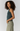 A woman with curly dark hair is shown in profile wearing a sleeveless ribbed knit top in a muted teal-green color, paired with light olive pants, against a solid light gray studio background.