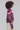 A side-profile view of a model wearing a cropped knit polo cardigan with horizontal stripes in purple, pink, and beige, paired with off-white pants against a solid light gray background.