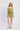 A woman modeling a light olive green two-piece set consisting of a tie-front smocked camisole top and matching smocked shorts, paired with woven tan flat shoes, against a plain white studio background.