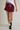 A person wearing a burgundy drawstring mini sweat skirt with white side stripes, styled with a white t-shirt, white socks, and black sneakers against a neutral studio background.
