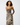 A woman modeling a sleeveless midi dress with a zebra print pattern in beige, black, and brown, set against a plain light gray studio background.