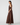 A woman modeling a floor-length brown satin gown with a draped cape-style back detail, shown in side profile against a light gray studio background.