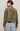 A woman shown from the back wearing an olive green cropped suede jacket and light blue jeans, photographed against a plain light beige studio background.