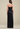 A woman shown from the back wearing a strapless black pleated maxi dress with a structured bodice and flowing skirt, photographed against a plain warm beige studio background.