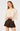 A woman modeling a cream long-sleeve blouse with lace trim and a bow at the neckline, paired with a dark brown pleated skirt, against a plain light gray studio background.