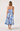 A woman viewed from behind wearing a sleeveless midi dress with thin straps and a blue floral print on a light background, paired with beige high heels, against a plain light gray studio backdrop.