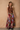 A woman stands against a textured beige wall wearing a brown draped halter top and a long multicolor patchwork-patterned skirt.