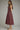 A woman modeling a sleeveless A-line midi dress in a deep burgundy color with a white polka dot pattern, shown from a side profile against a neutral gray studio background.