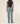 Rear view of a model wearing light-wash blue straight-leg jeans and a dark charcoal gray short-sleeve top against a plain off-white studio background.