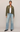 A model stands against a plain light beige studio background wearing an olive green cropped suede jacket over a white tank top, paired with light-wash wide-leg blue jeans and black boots.