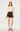 A model stands against a plain light gray studio background wearing a cream long-sleeve blouse with a ruffled Peter Pan collar and tie detail, paired with dark brown pleated mini shorts and dark brown high heels.