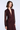 A woman modeling a long-sleeve, deep burgundy dress with a textured dotted pattern, a draped wrap-style bodice, and sheer mesh sleeves, set against a light gray studio background.