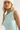 A close-up of a blonde model wearing a pale mint green ribbed tank top with sheer mesh side panels and a cream beanie against a plain light background.