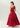 A woman modeling a vibrant red one-shoulder formal gown with a pleated bodice and full skirt, shown from a three-quarters back angle against a light gray studio background.