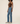 A model wearing a pair of high-waisted, straight-leg blue jeans, paired with a brown tank top and light gray sneakers, standing against a solid off-white studio background.