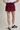 A person shown from the waist down wearing a burgundy athletic mini skirt with white side stripes, a white t-shirt, white socks, and black Adidas sneakers against a plain light gray studio background.