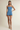 A model wearing a light blue denim romper with ruffled straps and hem, paired with beige heeled sandals, against a plain beige studio background.