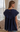 A woman viewed from the back wearing a navy blue short-sleeve top with a wide ruffled yoke and lace trim, standing in a living room setting with a sofa and curtains in the background.