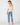 A woman is shown from the back wearing a cream-colored, semi-sheer puff-sleeve top with a scalloped neckline, paired with light-wash blue jeans and beige flat shoes, against a plain white studio background.
