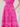A close-up of a bright pink midi dress with embroidered daisy-like floral motifs and a smocked waistband, shown on a model with one hand in the pocket.