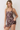 A woman modeling a brown spaghetti-strap mini dress with a botanical print featuring green leaves, beige stems, and pink flowers, set against a plain light gray background.