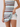 A close-up of a model wearing a sleeveless, bodycon mini dress with horizontal stripes in brown, cream, light blue, and white, layered over white fitted shorts against a plain light gray studio background.