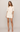 A woman modeling a short cream-colored dress with short sleeves, lace trim, and embroidered details on the bodice, paired with nude high-heeled sandals against a light gray studio background.
