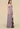 A woman modeling a strapless floor-length gown in a dusty lavender-purple color, featuring a ruched bodice and a pleated skirt with a large abstract floral print on the lower half, set against a solid beige background.