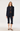 A woman models a dark navy blue two-piece suit consisting of a double-breasted blazer and matching cropped trousers, styled with a brown and beige striped knit top and brown pointed-toe heels against a plain light gray studio background.