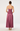 A woman viewed from the back wearing a sleeveless maxi dress with thin straps, a fitted lace-embroidered bodice, and a flowing satin-like skirt in a dusty rose or mauve tone, paired with gold high heels against a light gray studio background.