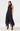A woman modeling a sleeveless high-neck midi dress with a navy blue base and small white polka dots, paired with brown suede ankle boots, against a plain white studio background.