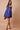A woman modeling a short blue spaghetti-strap dress with a gathered bow at the bust and a button placket, styled with brown sandals and a brown handbag against a solid beige studio backdrop.