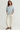 A woman modeling a light blue and white vertically striped button-down shirt with a peplum waist and rolled sleeves, paired with off-white wide-leg pants and dark brown pointed-toe heels, against a plain light gray studio background.