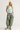 A woman modeling a light blue ribbed tank top, olive green baggy pants with a gray shirt tied around the waist, beige sneakers, and a cream beanie against a plain light gray studio background.