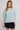 A woman modeling a light blue fuzzy short-sleeve knit top with a raw hem, paired with a black mini skirt, against a plain light gray studio background.