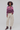 A model stands against a light gray studio background wearing a cropped, long-sleeve knit polo-style sweater with horizontal stripes in pink, purple, and beige, paired with off-white wide-leg cropped pants and dark brown wedge ankle boots.