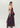 A woman modeling a strapless black maxi dress with a large magenta and purple floral print, shown from a three-quarters back view against a light gray studio background.