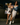 Two women posing together in a dark, stylish interior setting, wearing off-the-shoulder cream lace and satin dresses with denim jackets, white high-top sneakers, and sunglasses.