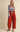 A woman modeling a cozy knit sweater with blue, orange, and white Fair Isle-style stripes, paired with high-waisted rust red corduroy pants and tan suede shoes, against a solid beige studio background.