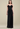 A woman modeling a strapless black evening gown with a ruffled sweetheart bodice and a floor-length pleated skirt, photographed against a plain warm beige studio background.