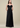 A woman modeling a strapless black pleated formal gown with a sweetheart neckline and ruffled shoulder details, photographed against a plain beige studio background.