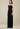 A woman modeling a strapless black pleated maxi gown with a structured bodice and ruffled shoulder detail, photographed against a plain beige studio background.