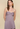 A woman modeling a strapless pleated dress in a muted dusty lavender color, set against a solid beige studio background.